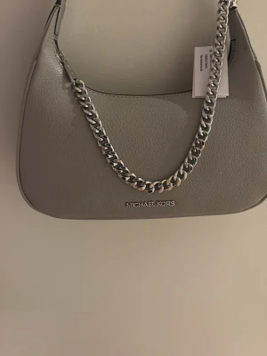 Grå axelväska från Michael Kors - Sprillans ny väska med lappen kvar och dessutom aldrig använd! Snygg grå handväska från Michael Kors i slätt läder med en chunky silverfärgad kedja som axelrem. Väskan har en stilren, avlång form och diskret logga framtill. Perfekt för dig som gillar minimalistisk och trendig design. Den är tidlös och fungerar i vått och torrt!