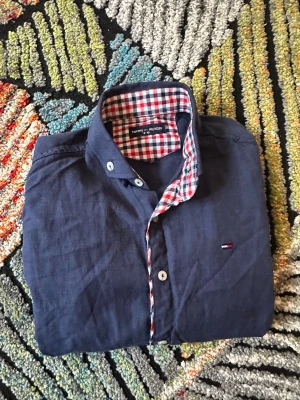 Marinblå skjorta från Tommy Hilfiger - Snygg marinblå skjorta från Tommy Hilfiger med rutigt foder i krage och ärmslut. Skjortan har långa ärmar, knappar framtill och liten broderad logga på bröstet. Perfekt för dig som gillar klassisk stil med en twist.