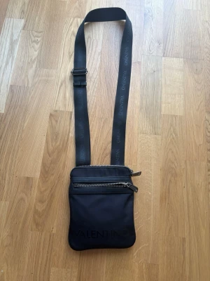 Svart axelväska från Valentino - Snygg svart axelväska från Valentino med justerbart axelband där loggan är tryckt. Väskan har två dragkedjefack framtill och är tillverkad i ett slätt, tåligt material. Perfekt för att bära det viktigaste med stil.