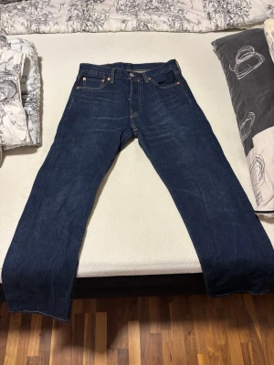 Mörkblå Levi's 501 jeans - Säljer ett par klassiska mörkblå jeans från Levi's med raka ben och fem fickor. Jeansen har gylf med knappar och tydliga kontrastsömmar. Tillverkade i slitstark denim, perfekt för en avslappnad och tidlös look. Jeansen använda endast 1 gång.