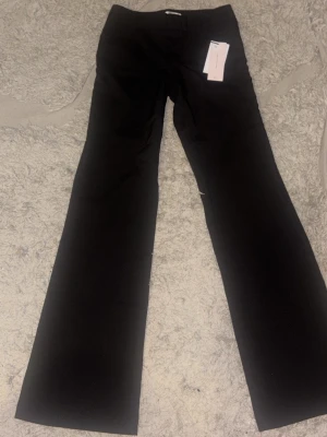 Svarta utsvängda kostymbyxor - Snygga svarta kostymbyxor med utsvängda ben och hög midja. Byxorna har fickor med lock och knappdetaljer baktill. Perfekta för en stilren och trendig look. Materialet ser ut att vara en mjuk och följsam mix, troligen polyester eller viskos. Dem är ganska långa i längden 