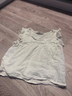 Vit blus med volang från H&M - Söt vit blus från H&M med volangdetaljer vid axlarna och diskret broderat mönster i tyget. Blusen är ärmlös och har en lätt och luftig känsla, perfekt för varmare dagar.