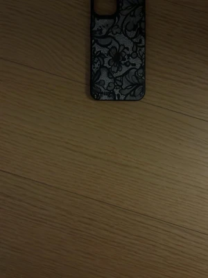 Svart mobilskal med spetsmönster iPhone 13 - Snyggt svart mobilskal med detaljerat spetsmönster på baksidan. Skalets material känns som tålig plast och det har en öppning för kameran. Passar dig som vill ha ett elegant och unikt skydd till mobilen. till iPhone 13