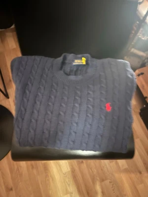 Mörkblå kabelstickad tröja Polo - Kabelstickad mörkblå tröja från Polo Ralph Lauren med rund halsringning och klassisk röd broderad logga på bröstet. Tröjan har ribbade muddar och är perfekt för en stilren och tidlös look.