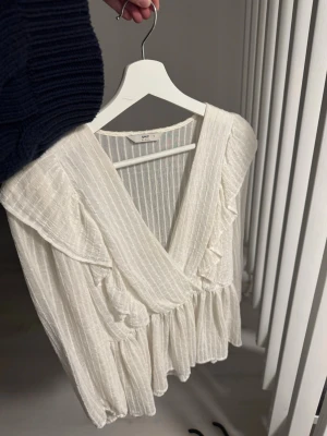 Vit omlottblus med volanger från ONLY - Superfin vit omlottblus från ONLY med volangdetaljer och diskret randigt strukturmönster. Blusen har v-ringning och långa ärmar, samt en luftig och flowy passform. Perfekt att styla med jeans eller kjol för en trendig look.