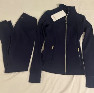 Marinblå träningsset från Lululemon - Snyggt marinblått träningsset från Lululemon med en figurnära jacka med guldfärgade dragkedjor och två fickor framtill. Tillhörande tights i samma färg och material. Perfekt för gymmet eller löprundan. Storlek 2