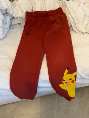 Röda Pokémon mjukisbyxor Pikachu - Säljer ett par röda mjukisbyxor med Pikachu-tryck på ena benet och elastisk midja. Byxorna har resår vid bensluten och är perfekta för dig som älskar Pokémon. Materialet är mjukt och bekvämt, perfekt för chill dagar hemma.