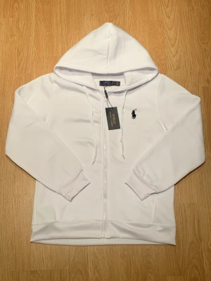 Vit hoodie från Polo Ralph Lauren - Snygg vit hoodie från Polo Ralph Lauren med dragkedja framtill och klassisk svart broderad logga på bröstet. Hoodien har huva med dragsko, ribbade muddar och är tillverkad i mjukt bomullsmaterial. Perfekt för en clean och stilren look.