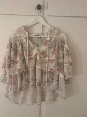 Blommig volangblus med knytband - Romantisk blus i transparent, lätt material med blommigt mönster i rosa och grönt. Blusen har volanger, v-ringning med knytband och långa ärmar. Perfekt för dig som gillar en bohemisk och söt stil.