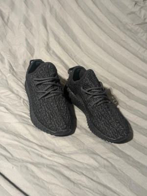 Svarta Yeezy Boost 350 sneakers - Säljer ett par svarta Yeezy Boost 350 sneakers med stickad ovandel och mönstrad textur. Skorna har svarta snören, svart sula och en diskret röd detalj på hälen. Perfekta för dig som gillar streetwear och vill ha en ikonisk sneaker i garderoben.