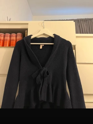 Mörkblå stickad tröja från Nelly - Mysig mörkblå stickad tröja från Nelly med knyt framtill och volangdetalj vid nederkanten. Tröjan har lång ärm och en djup v-ringning, perfekt för lager-på-lager-stil.