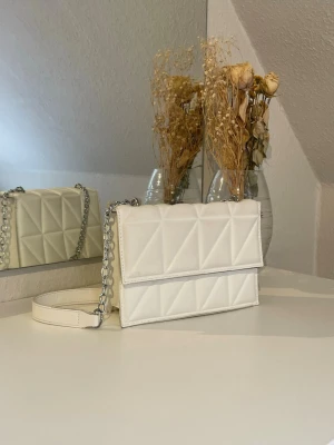 Vit quiltad handväska med kedja - Säljer en stilren vit handväska i quiltat konstläder med geometriskt mönster. Väskan har ett lock med magnetlås och en axelrem i kombinerat konstläder och silverfärgad kedja.💕