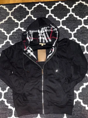 , - Säljer en svart zip-hoodie från Burberry med klassiskt rutigt foder i huvan och dragsko. Hood Autentiskt Burberry-bro studio på bröstet. Mjuk bomullskänsla och två fickor framtill. Helt ny använd 1 gång 