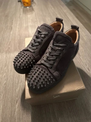 Mörkgrå sneakers med nitar - Unika mörkgrå sneakers från Christian Louboutin med matta nitar över tån och ovandel i mocka. Skorna har klassisk snörning, låg profil och en lyxig känsla med beige läderfoder. Perfekta för dig som vill sticka ut med din stil.