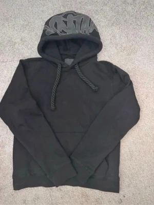 Svart hoodie från Syna world - Svart hoodie från Sefyr Sankt med tryckt logga på etiketten och texten '0% FUCKERIES'. Klassisk modell med huva och ribbade muddar. Mjuk insida som känns skön mot huden. Perfekt för en avslappnad streetwear-look.