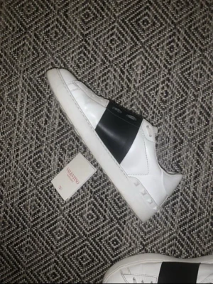 Valentino vita sneakers med svart band - Snygga vita sneakers från Valentino med ett brett svart band över mitten. Skorna har klassisk låg profil och är tillverkade i läder med detaljer som nitar på hälen. Perfekt för dig som vill ha en stilren och exklusiv look. Självklart äkta som man ser tydligt. Strlk 42,5 men passar 43 och 42