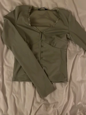 Ljusgrön långärmad topp med knappar - Supersöt ljusgrön topp med långa ärmar och knäppning framtill. Toppen har en v-ringad hals med spetsdetaljer och figurnära passform. Perfekt att styla med jeans eller kjol för en trendig look.