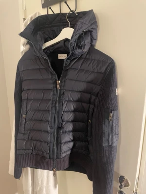 Mörkblå pufferjacka från Moncler - Snygg mörkblå pufferjacka från Moncler med stickade ärmar och huva. Jackan har dragkedja framtill, två fickor med tryckknappar och en extra ficka med dragkedja på ärmen. Moncler-logga på vänster ärm. Perfekt för kalla vinterdagar.