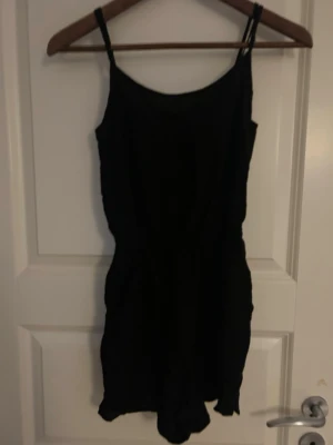 Svart byxdress från Divided strl 32 - Svart byxdress från Divided i storlek 32. Modellen har smala axelband med knyt och resår i midjan för en skön passform. Materialet är mjukt och lätt bomull, perfekt för varma dagar. Enkel och stilren design med korta ben och omlott-detalj upptill.