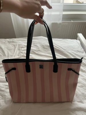 Rosa och vitrandig handväska - Säljer en snygg handväska med rosa och vita ränder, svarta handtag och detaljer. Väskan har en praktisk dragkedja på baksidan och ett litet emblem framtill. Perfekt för dig som gillar en färgglad och trendig look.