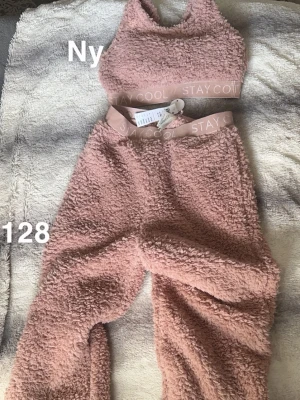 Rosa fluffigt loungewear-set  - Mysigt loungewear-set i fluffigt teddy-material från Lindex. Setet består av ett rosa top med bred resår och matchande byxor med hög midja och elastisk linning. Perfekt för chill dagar hemma. Materialet är supermjukt och ger en cozy vibe.🌸 Ny med lapp. Storlek 128