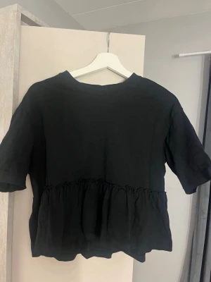 Svart volangtopp med kort ärm - Svart topp med volang nedtill och korta ärmar. Modellen är loose fit och har rund halsringning. Perfekt att styla med jeans eller kjol för en trendig look.