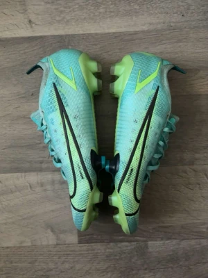 Nike Mercurial blågröna fotbollsskor - Säljer ett par Nike Mercurial fotbollsskor i blågrönt med svarta och neongula detaljer. Skorna har snörning och mönstrad ovandel för extra grepp på planen. Sulan är gjord för spel på gräs och har fasta dobbar. Perfekta för dig som vill sticka ut på fotbollsplanen!
