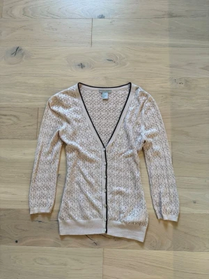 Beige mönstrad kofta med v-ringning - Snygg rosa kofta med svart kant och knappar framtill. Koftan har ett diskret mönster över hela plagget och djup v-ringning. Lätt och tunn i materialet, perfekt för lager-på-lager. Långa ärmar och ribbade muddar.