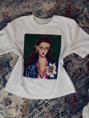 Vit t-shirt med konstmotiv - Vit t-shirt med ett stort färgstarkt porträtt på framsidan som föreställer en person med långt hår, blommor och detaljerad klädsel. T-shirten har rund hals och korta ärmar. Det är en väldigt cool och unik T-shirt som passar till mycket! Den är cool och något alldeles extra!🪻✨