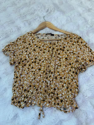 Blommig gul blus från LCW Casual - Söt blommig blus i gult från LCW Casual med korta ärmar och rund hals. Blusen har små vita och svarta blommor, knappar framtill och en dragsko i midjan för en snygg passform. Tillverkad i ett lätt och luftigt tyg, perfekt för varma dagar.