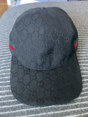 Svart Gucci keps - Säljer en svart keps från Gucci med det klassiska GG-monogrammet över hela kepsen. På sidorna finns gröna och röda ränder som ger en ikonisk touch. Kepsen har böjd skärm och är tillverkad i ett slitstarkt tygmaterial. Perfekt för dig som vill ha en stilren och lyxig accessoar.
