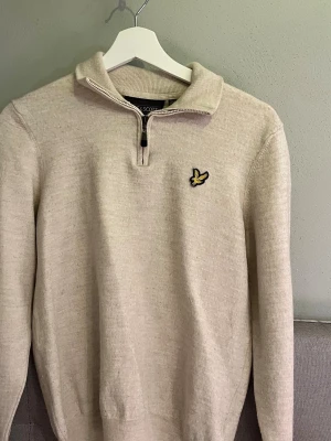 Beige stickad tröja från Lyle & Scott - Beige stickad tröja från Lyle & Scott med halv dragkedja och klassisk gul logga på bröstet. Tröjan har lång ärm, ribbade muddar och krage. Perfekt för lager-på-lager och enkel att matcha med jeans eller chinos.