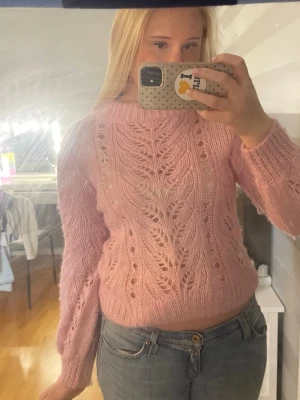 Rosa stickad tröja med hålmönster - Säljer en fluffig rosa stickad tröja med vackert hålmönster och ribbade muddar. Tröjan har rund hals och långa ärmar, perfekt för att ge en mysig och trendig look. Materialet ser ut att vara mjukt och lätt, troligen mohair eller liknande.