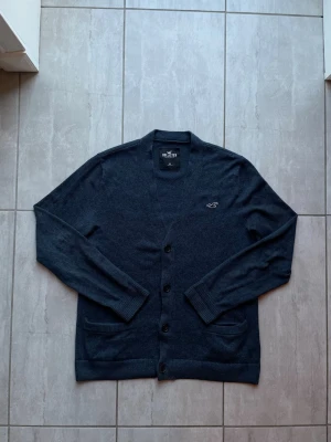 Mörkblå cardigan från Hollister - Stilren mörkblå cardigan från Hollister med knäppning framtill och två fickor. Cardigan har ribbade muddar vid ärmslut och nederkant samt en liten broderad logga på bröstet. Perfekt för lager på lager och enkel att matcha.