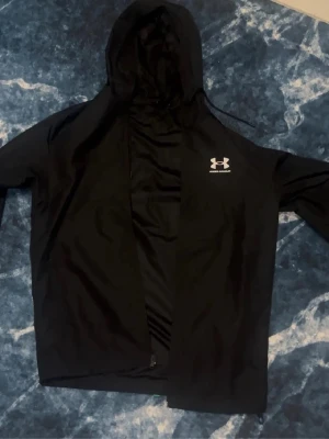 Svart vindjacka från Under Armour - Svart vindjacka från Under Armour med huva och dragkedja framtill. Jackan har en stilren design med logga på bröstet och justerbar huva. Perfekt för blåsiga dagar och enkel att matcha med olika outfits.