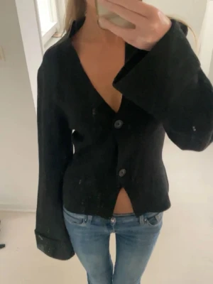 Svart kofta med vida ärmar Ginatricot - Svart kofta från Ginatricot med två knappar framtill och djup v-ringning. Modellen har vida ärmar med dekorativa knappar vid ärmsluten och en något kortare passform. Perfekt att styla med jeans eller kjol för en trendig look. Nästan helt ny, använd några gånger