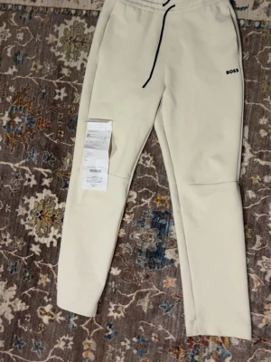 Beige mjukisbyxor från BOSS - Snygga beige mjukisbyxor från BOSS med svart dragsko i midjan och diskret logga på vänster höft. Byxorna har en slim fit-modell och är tillverkade i mjukt material som känns skönt mot huden. Perfekta för en chill och stilren look.