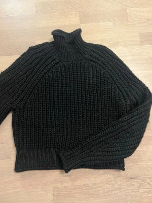 Svart grovstickad polotröja - Säljer en svart grovstickad polotröja med hög krage och långa ärmar. Tröjan har en chunky känsla och är perfekt för kyliga dagar. Materialet känns mjukt och mysigt, och modellen är något boxig med ribbstickade muddar. Aldrig använd 💞