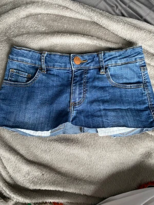 Blå jeanskjol med slitningar - KOLLA VERKLIGEN MÅTTEN INNAN KÖP! Snygga blå jeansshorts med rå kant och slitna detaljer på bakfickorna. Klassisk femficksmodell med knapp och dragkedja framtill. Perfekta för sommaren och ger en avslappnad vibe. Den är superrrr kort så man vet. Kolla måtten på bilderna. Själv är jag ca 162 för referens och är 14. Skriv om frågor och pris kan förhandlas!💗inga returer❌ fraktar direkt efter köp!📦