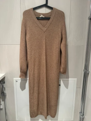 Beige stickad klänning med v-ringning - Mysig beige stickad klänning med v-ringad hals och långa ärmar. Klänningen har en rak passform och ribbstickad struktur som ger en avslappnad men ändå stilren look. Perfekt för lager på lager och enkel att styla med boots eller sneakers.