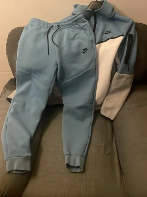 Nike Tech Fleece set blå/grå - Nike Tech Fleece set med ziptröja och byxor i ljusblått, vitt och grått. Tröjan har dragkedja, hög krage och svart Nike-logga på bröstet samt ficka på ärmen. Byxorna har resår i midjan och svart Nike-logga. Perfekt för chill eller träning.