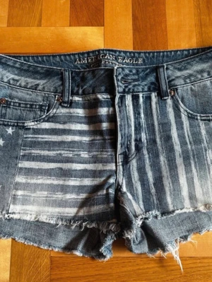 American Eagle jeansshorts med flaggmönster - Snygga jeansshorts från American Eagle med amerikanskt flaggmönster framtill och stjärnor på bakfickorna. Shortsen är blå, har råa kanter och klassisk femficksdesign. Perfekta för sommaren och ger en cool, avslappnad vibe.