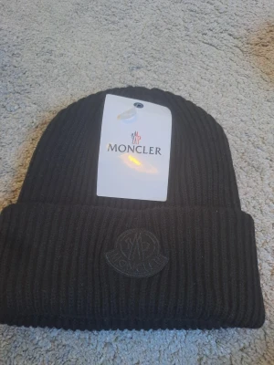 Svart Moncler mössa i ribbstickad ull - Säljer en svart ribbstickad mössa från Moncler med uppvikt kant och diskret Moncler-logga framtill. Mössan är tillverkad i mjuk ull och passar perfekt för kyliga dagar. Levereras med originalpåse och tag.