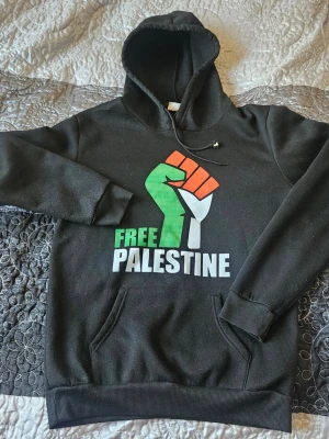 Svart hoodie Free Palestine tryck - Svart hoodie med huva och känguruficka framtill. På bröstet finns ett stort tryck med texten 'FREE PALESTINE' och en knuten näve i grönt, vitt och rött. Mjuk insida och ribbade muddar vid ärmslut och nederkant. Perfekt för dig som vill göra ett statement.