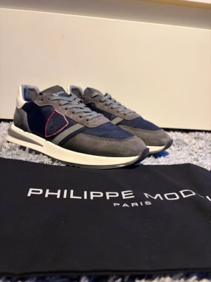 Philippe Model grå/blå sneakers - Snygga sneakers från Philippe Model i grått och marinblått med vita detaljer och en röd kant på sidan. Skorna har en klassisk låg siluett, mesh och mocka-paneler samt vita sulor. Perfekta för dig som gillar stilrena och trendiga sneakers. Perfekt nu till våren sommaren 