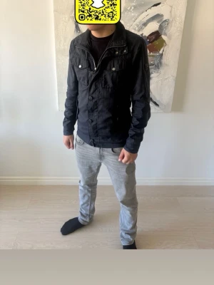 Marinblå Field Jacket från J.lindberg - Snygg svart Field Jacket från J.Lindberg med klassisk krage, knäppning framtill och två bröstfickor med lock. Jackan har en stilren patch på ena ärmen och är tillverkad i jeansmaterial. Perfekt för dig som gillar en clean och modern look.