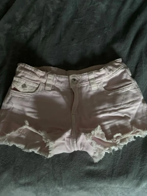 Ljusrosa jeansshorts med fransar - Säljer ett par ljusrosa jeansshorts med fransig kant och klassiska fickor fram och bak. Shortsen har bälteshällor, knapp och dragkedja framtill. Perfekta för varma sommardagar och en avslappnad stil.