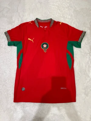 Marocko fotbollströja Puma röd L - Säljer en röd Marocko fotbollströja från Puma med gröna och guldiga detaljer. Tröjan har broderat landslagsemblem på bröstet, mönstrad krage och ärmslut samt DryCell-material som andas. Perfekt för dig som älskar fotboll och vill visa ditt stöd för Marocko.