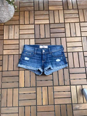 Blå jeansshorts med slitningar - Snygga blå jeansshorts från Hollister med slitna detaljer och fransig kant. Klassisk femficksmodell med låg midja och knappgylf. Perfekta för varma dagar och en avslappnad stil. Midjemått 37cm❣️