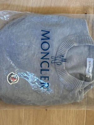 Grå Moncler tröja med logga - Snygg grå tröja från Moncler med blå logotyp tryckt på framsidan och klassisk Moncler-patch på ena sidan. Tröjan har rund halsringning och ribbade muddar vid ärmslut och nederkant. Perfekt för en stilren och avslappnad look.